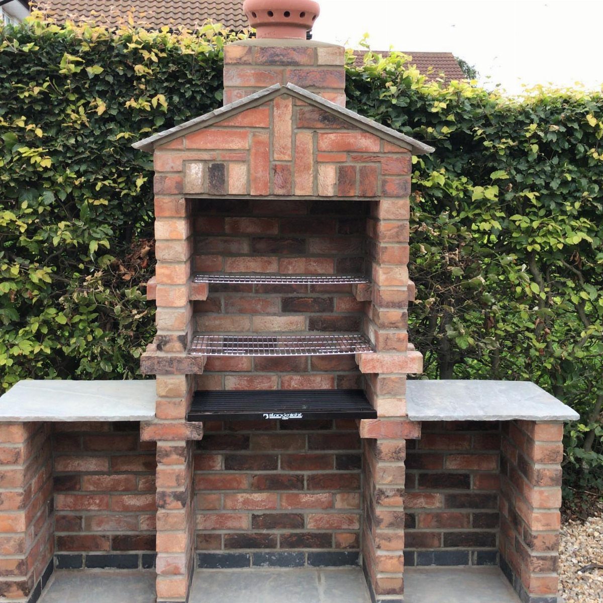 A brick outdoor barbecue with a chimney, metal grates, and stone countertops against a hedge.