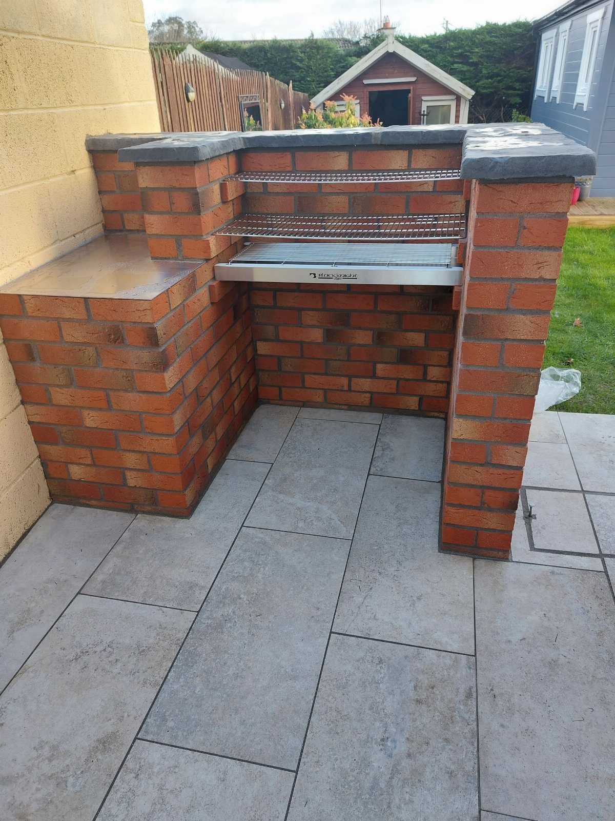 A red brick built-in barbecue with metal grates on a grey tiled patio.