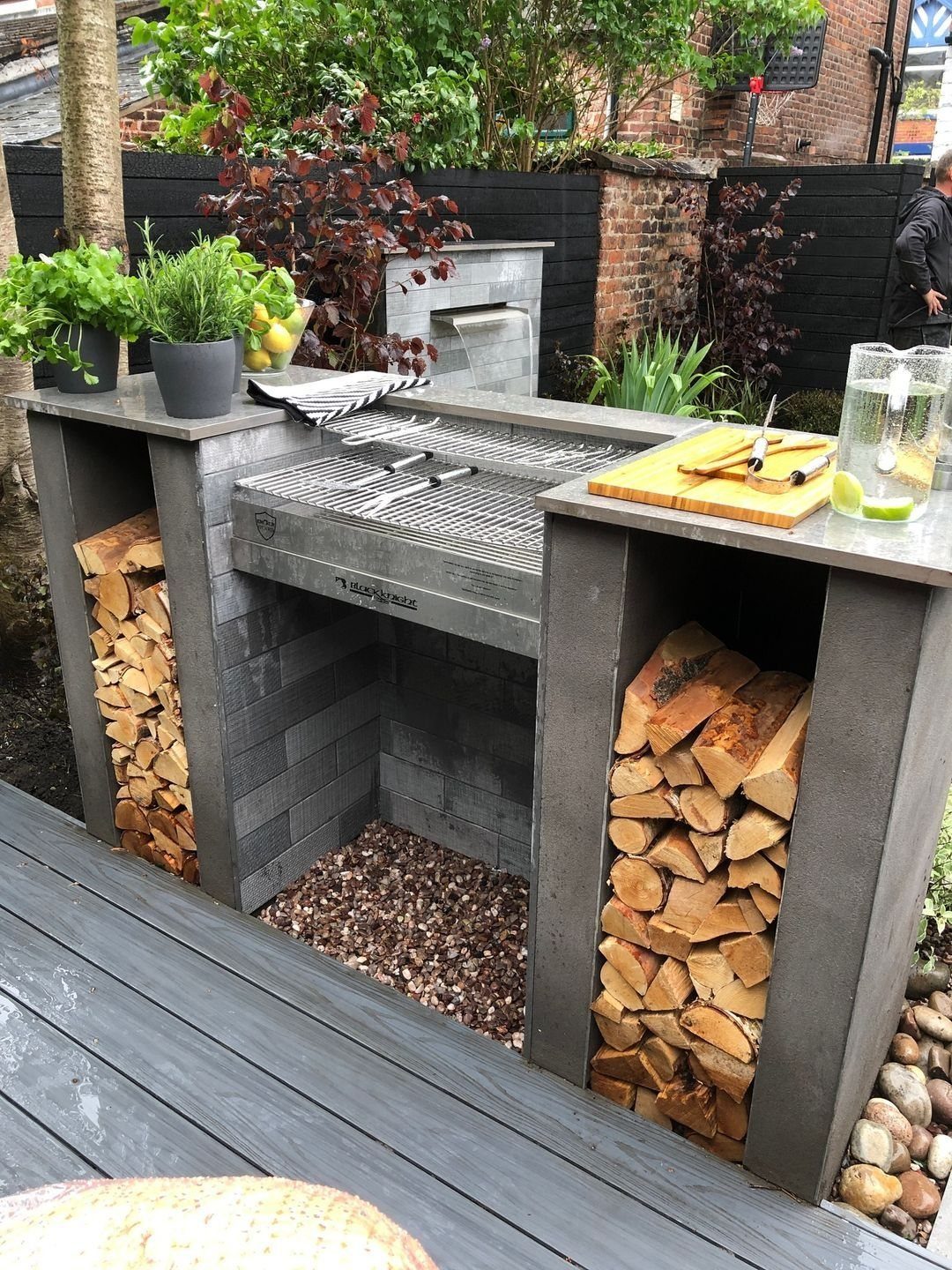 A modern dark grey minimalist outdoor grill station with stacked firewood in side compartments.