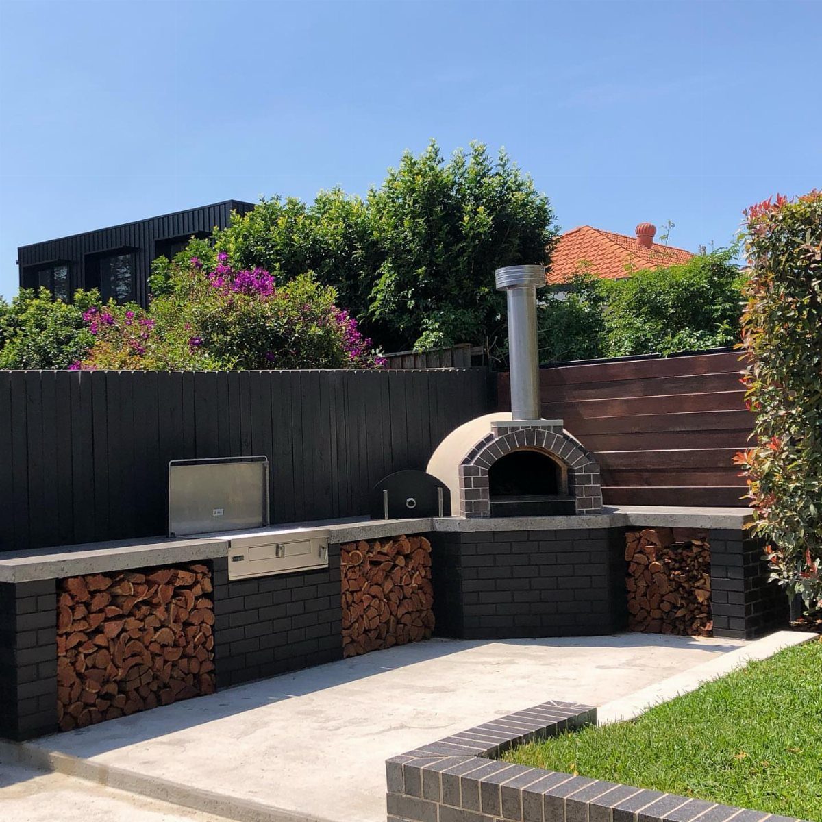 A dark brick outdoor kitchen with a pizza oven, grill, and stacked firewood.