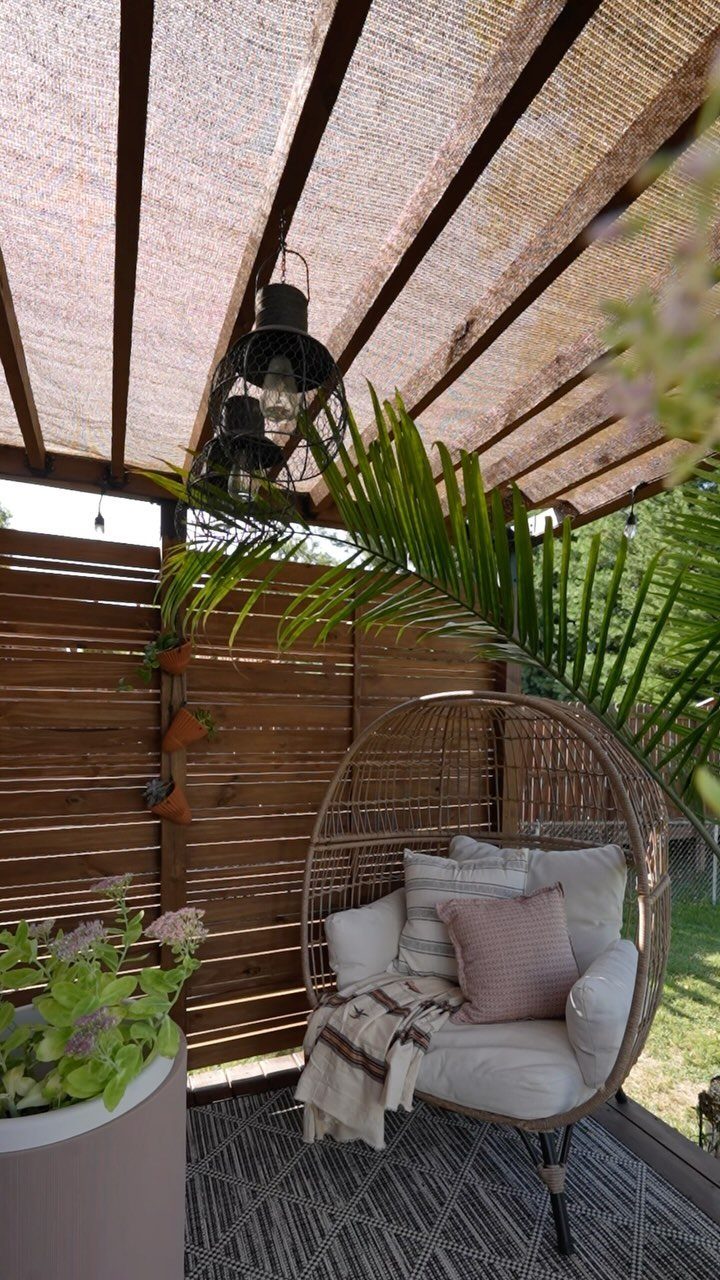 A woven sun shade canopy attached to a wooden pergola over a patio with a wicker egg chair.
