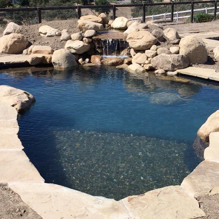 A stone-lined backyard swimming pool featuring a small waterfall and large natural rocks.
