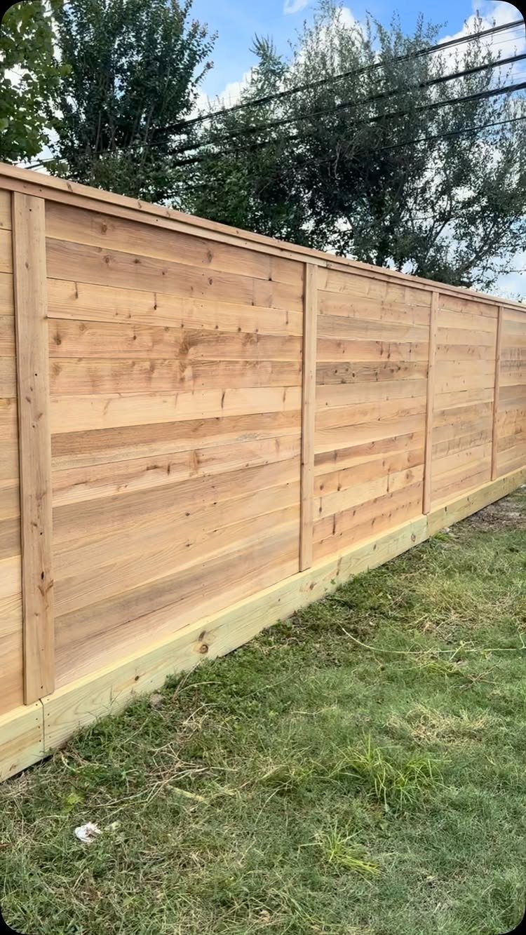 A long horizontal wooden privacy fence installed along a green grass lawn with trees in the background.