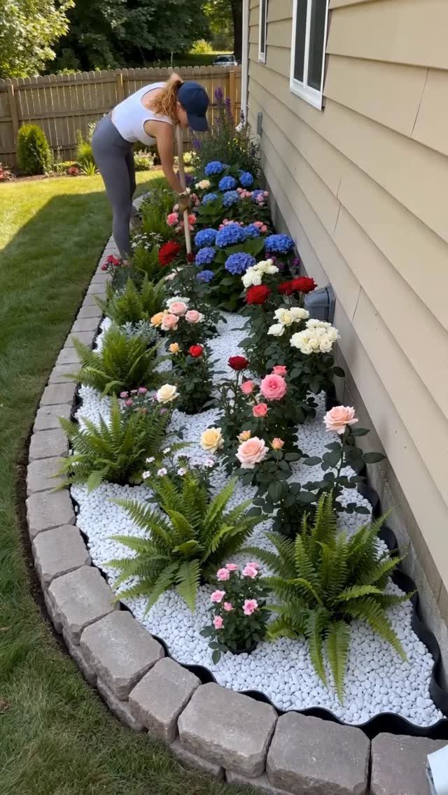 A person gardening in a flower bed with roses and ferns along a garage wall.