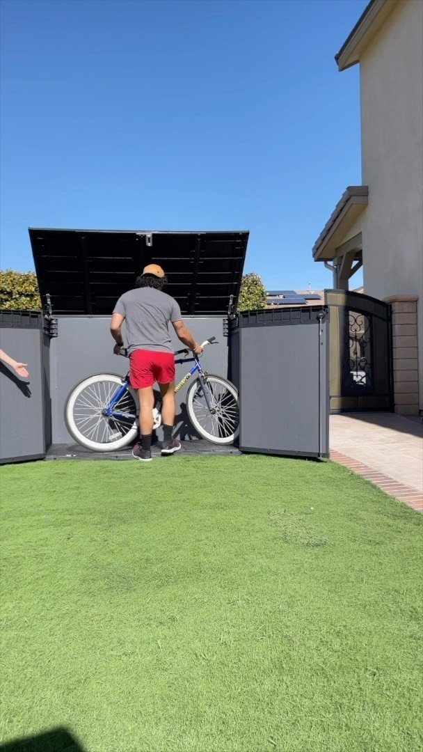 A person placing a blue bicycle into a large grey outdoor storage box on a lawn.