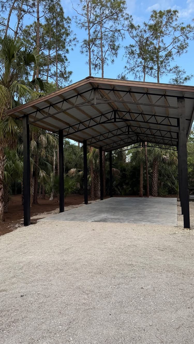 A large open-sided metal pole barn structure with a concrete floor surrounded by tropical trees.