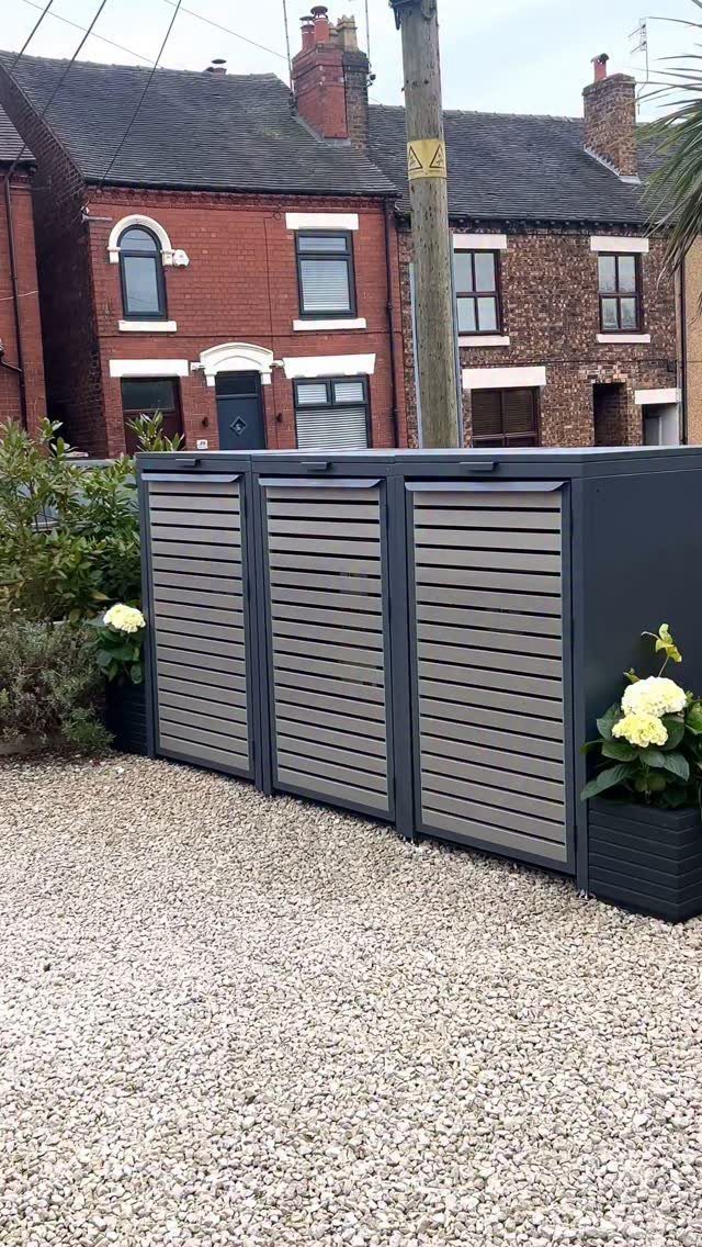 Dark grey outdoor storage unit with wooden slats sitting on a gravel patio near brick houses.