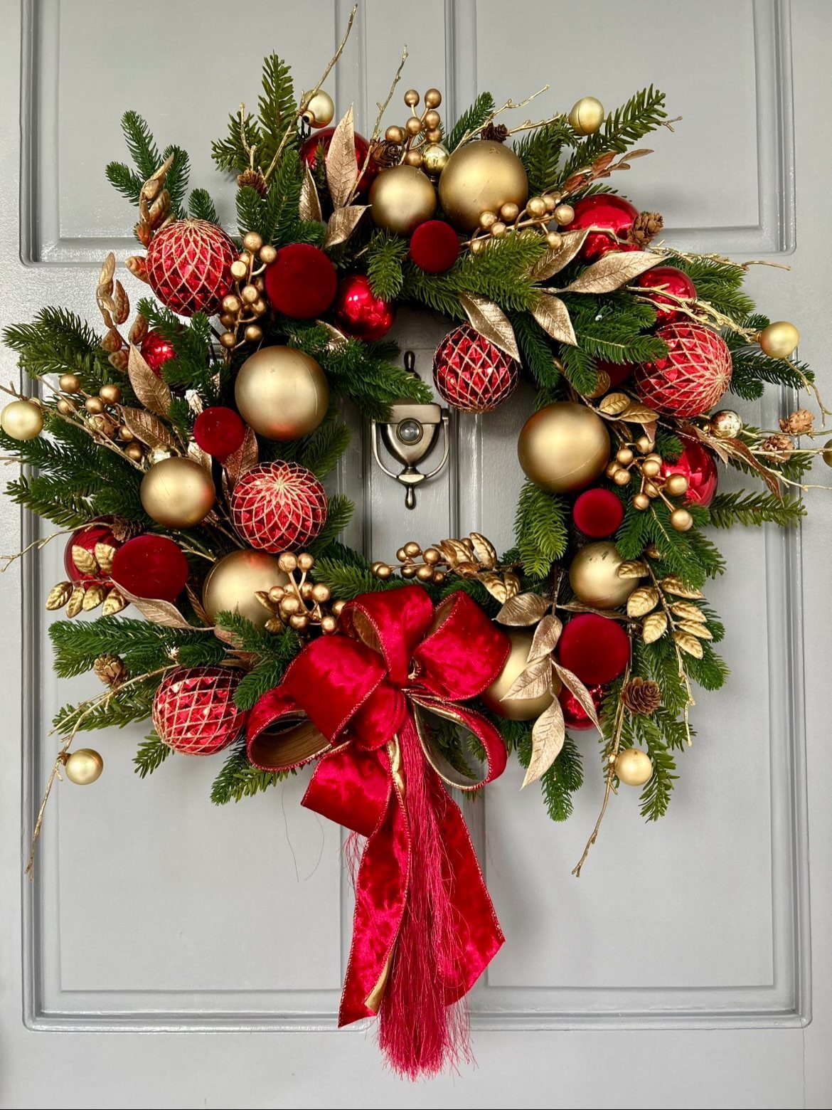 A festive evergreen wreath decorated with red and gold ornaments and a large red bow on a grey door.