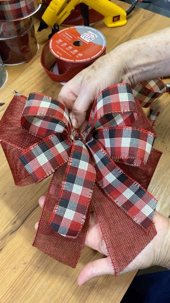 Hands holding a large handmade bow made of red and plaid layered ribbons on a wooden table.