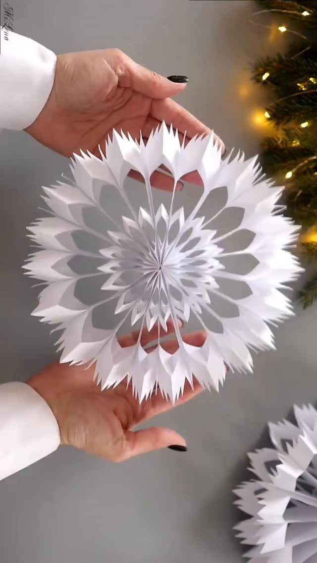 Hands holding a large, intricate white 3D paper snowflake decoration against a blurred Christmas tree background.
