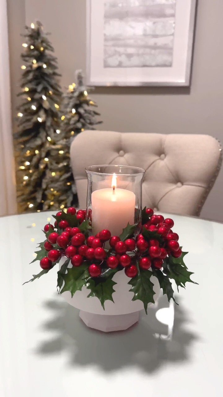 27 Christmas Display Ideas for Every Room of the Home 12 A lit candle in a glass vase surrounded by red berries and holly leaves on a white table.
