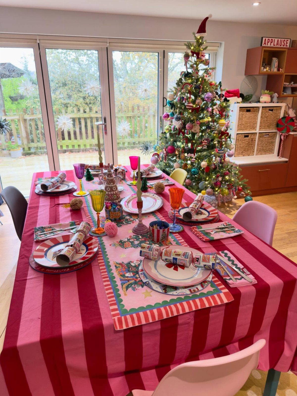 27 Christmas Display Ideas for Every Room of the Home 25 A festive dining table set with colorful plates, glassware, and crackers under a decorated Christmas tree.