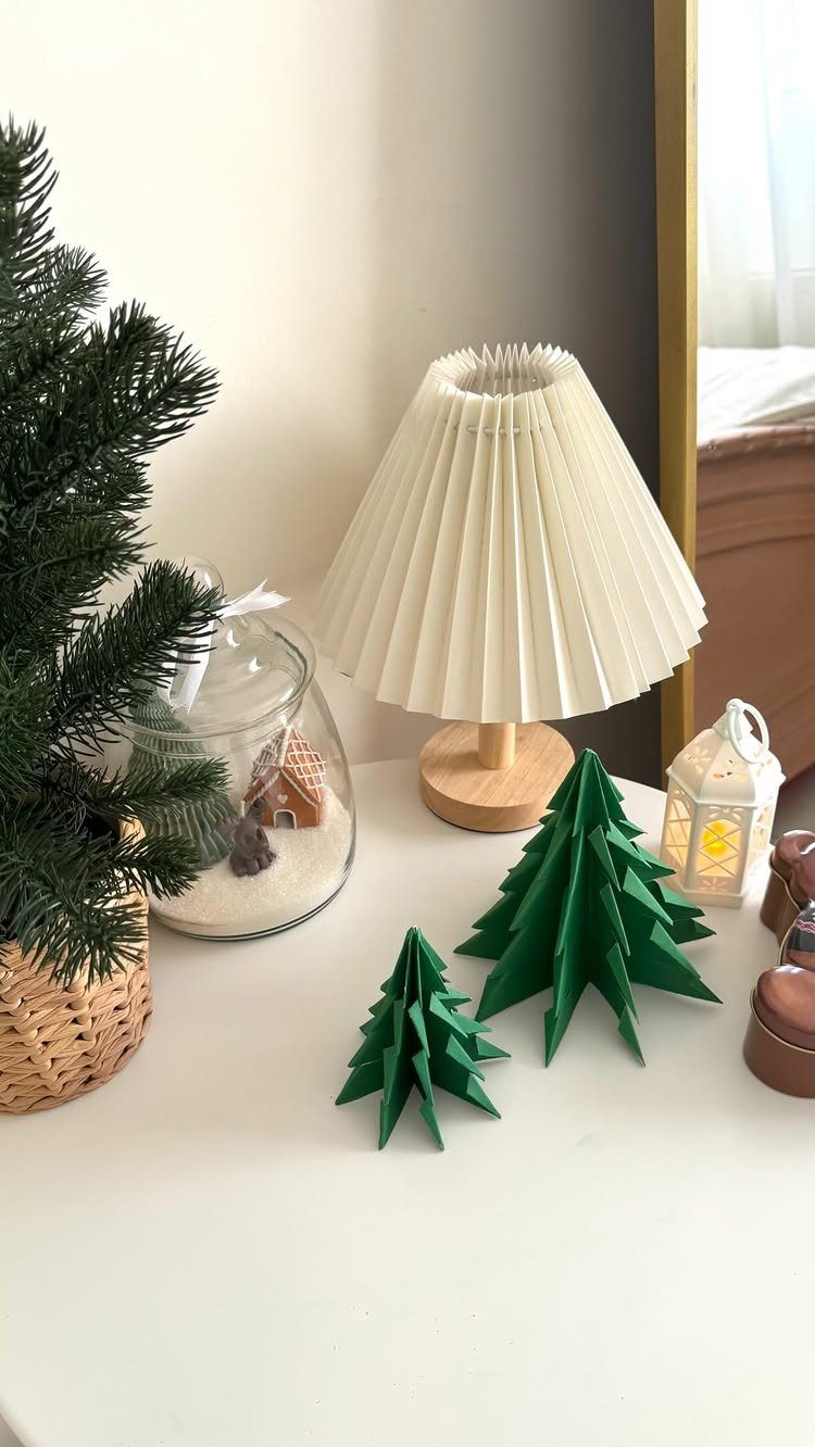Two small green geometric paper trees sitting on a white tabletop next to a lamp.