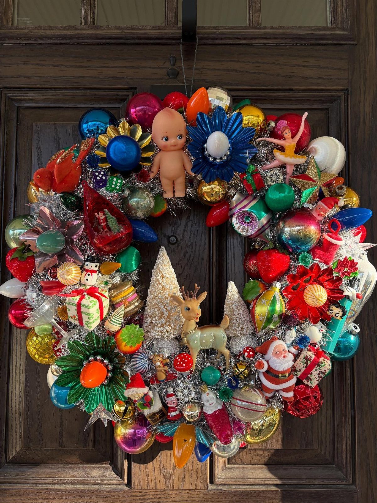 A colorful Christmas wreath decorated with ornaments and tinsel hanging on a dark wooden door.