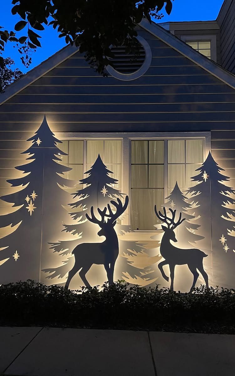 White silhouette cutouts of pine trees and deer glowing against a lit window at night.