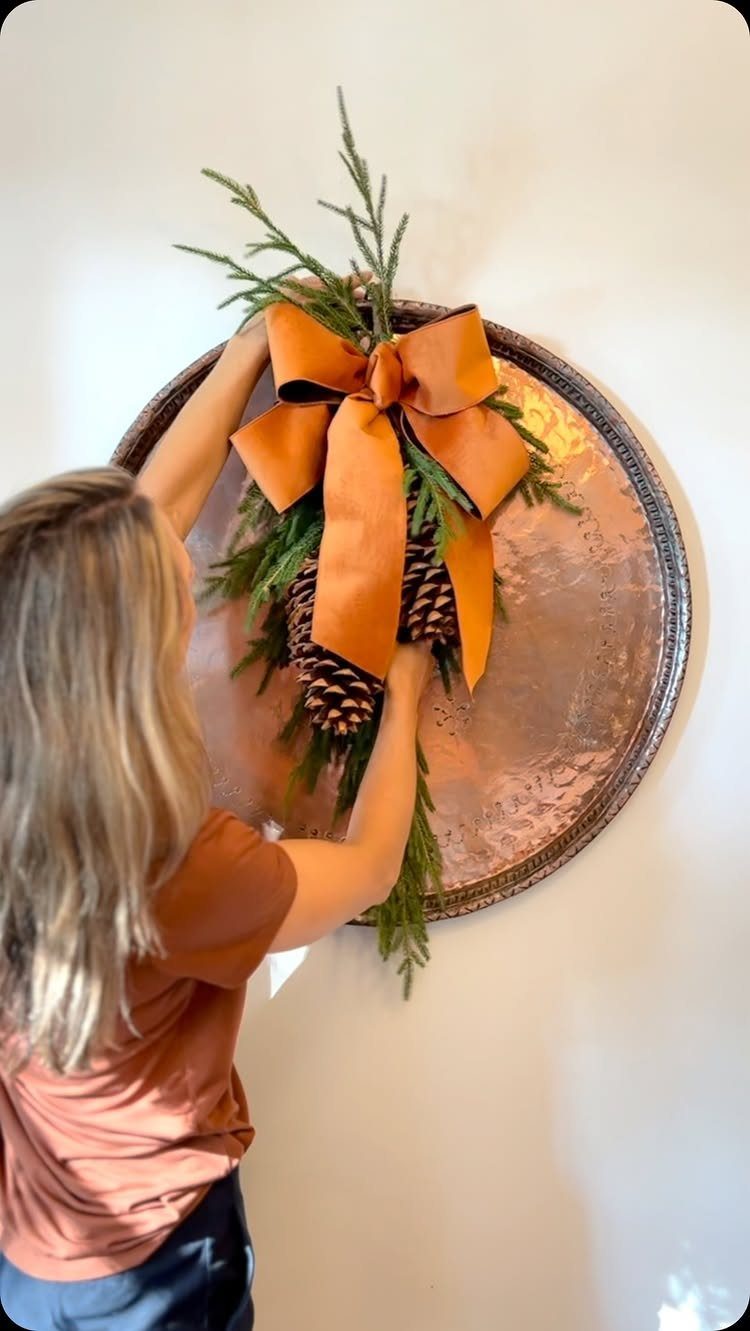 Christmas Wall Decor Ideas: 9 Ways to Dress Vertical Space 9 Woman hanging a copper plate decorated with evergreen branches, pinecones, and an orange bow on a wall.