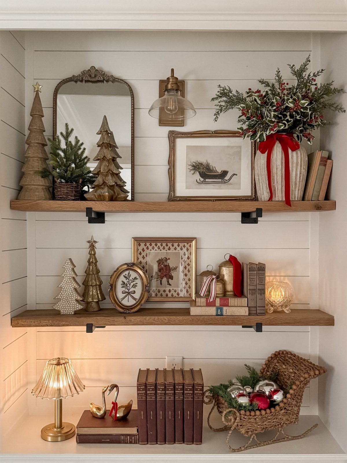 Christmas Wall Decor Ideas: 9 Ways to Dress Vertical Space 5 Wooden shelves decorated with miniature trees, framed holiday art, books, and greenery against white shiplap.