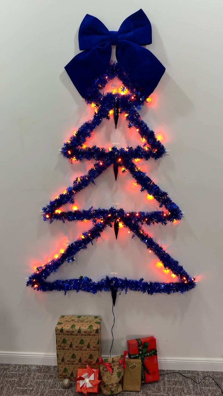 Christmas Wall Decor Ideas: 9 Ways to Dress Vertical Space 7 Blue tinsel Christmas tree outline with orange lights and a large blue bow on a white wall.