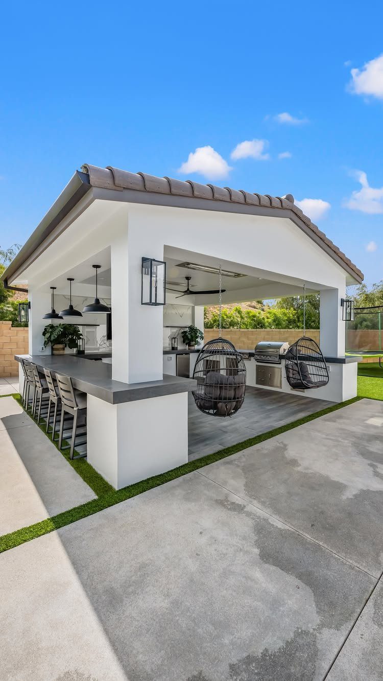 Coastal Outdoor Kitchen and 7 Ideas for Beachside Cooking Spaces 4 Modern white outdoor kitchen with a long bar, high stools, and hanging egg chairs under a roof.