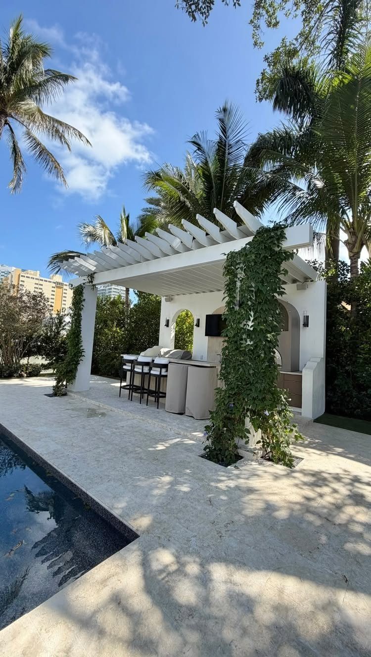 Coastal Outdoor Kitchen and 7 Ideas for Beachside Cooking Spaces 2 A white pergola with climbing vines shelters an outdoor kitchen and bar area by a pool.