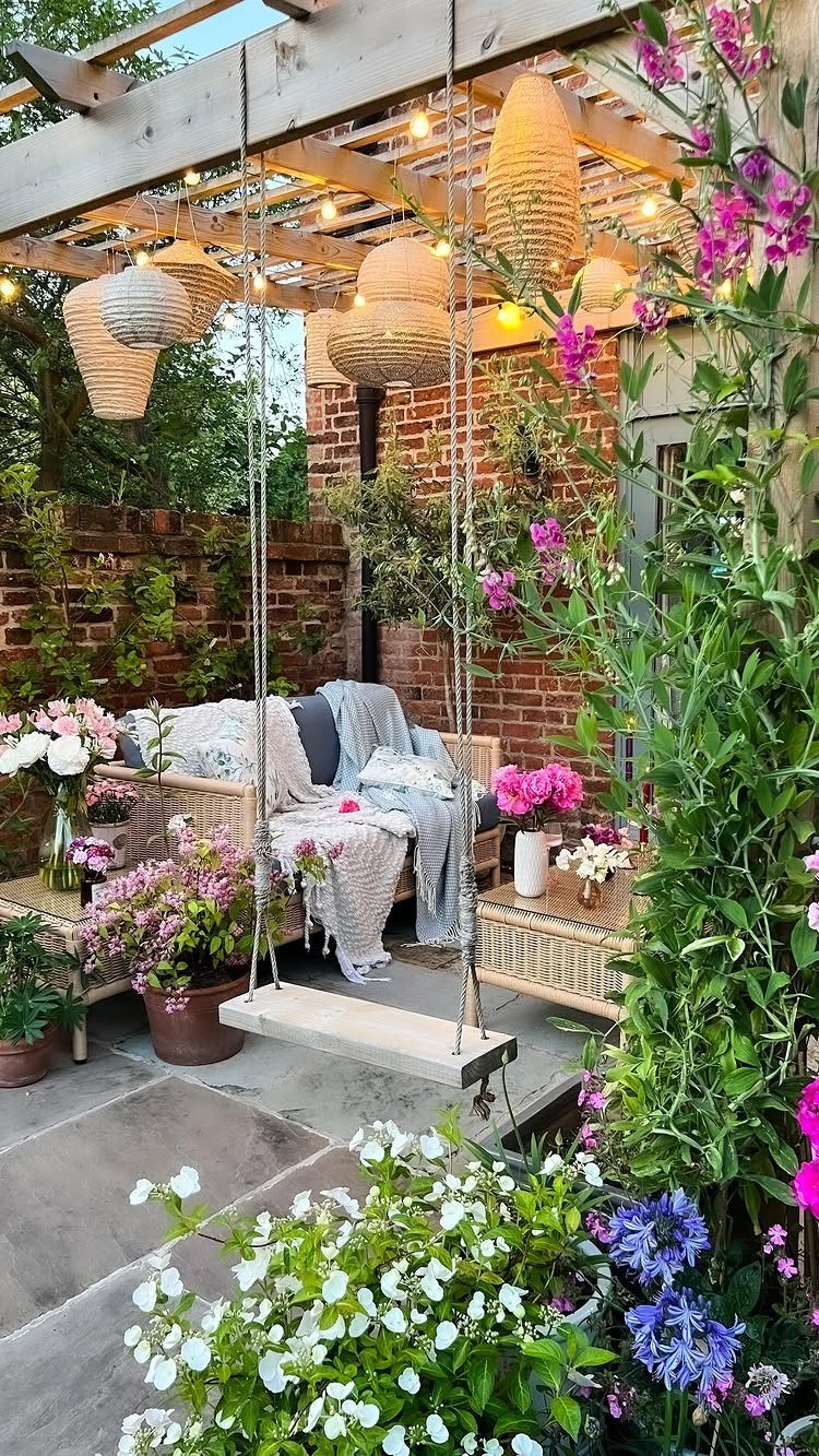 A cozy patio with a hanging swing seat, blooming flowers, and woven pendant lights under a pergola.