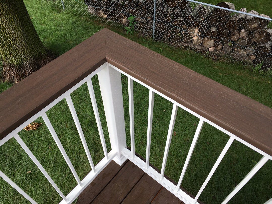 A wide brown wooden deck railing with white balusters overlooking a green lawn.