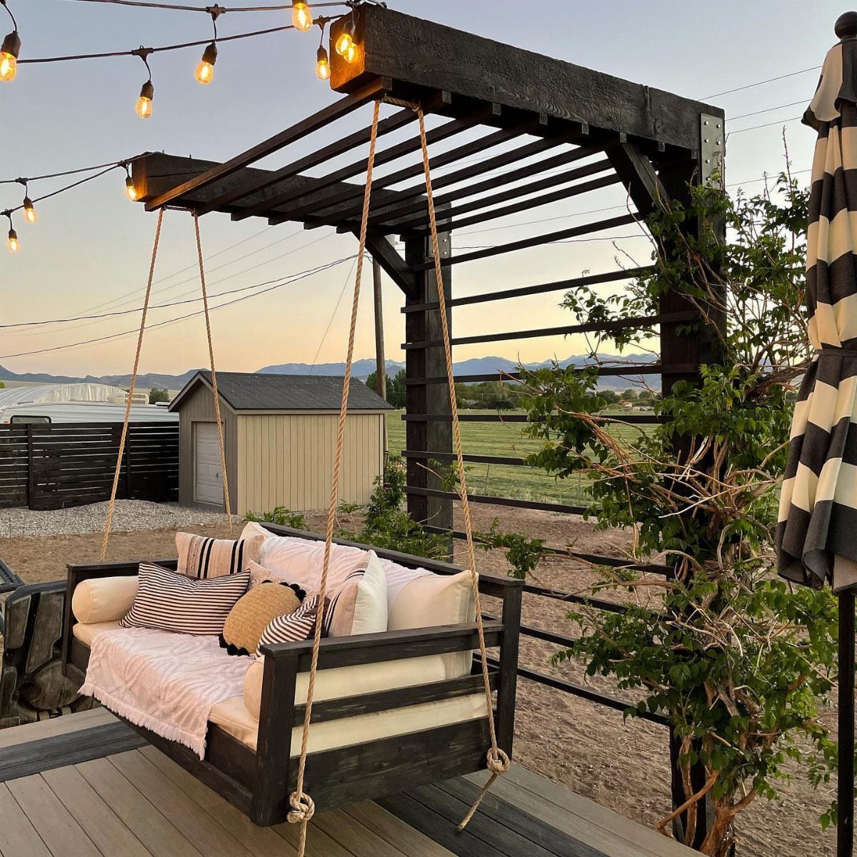 A dark wooden swing bed with white cushions hanging from a pergola on a deck.