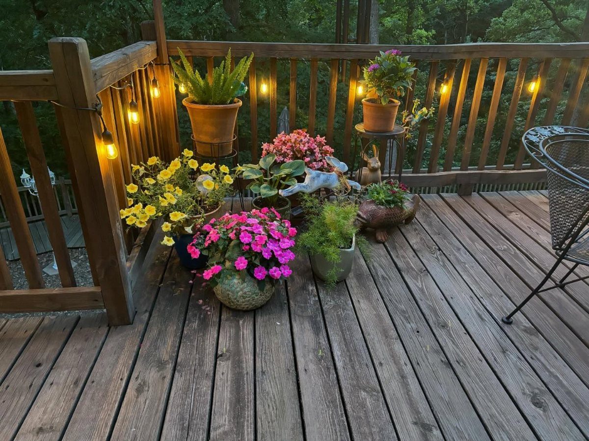 Colorful potted flowers and green plants arranged on a wooden deck with glowing string lights.