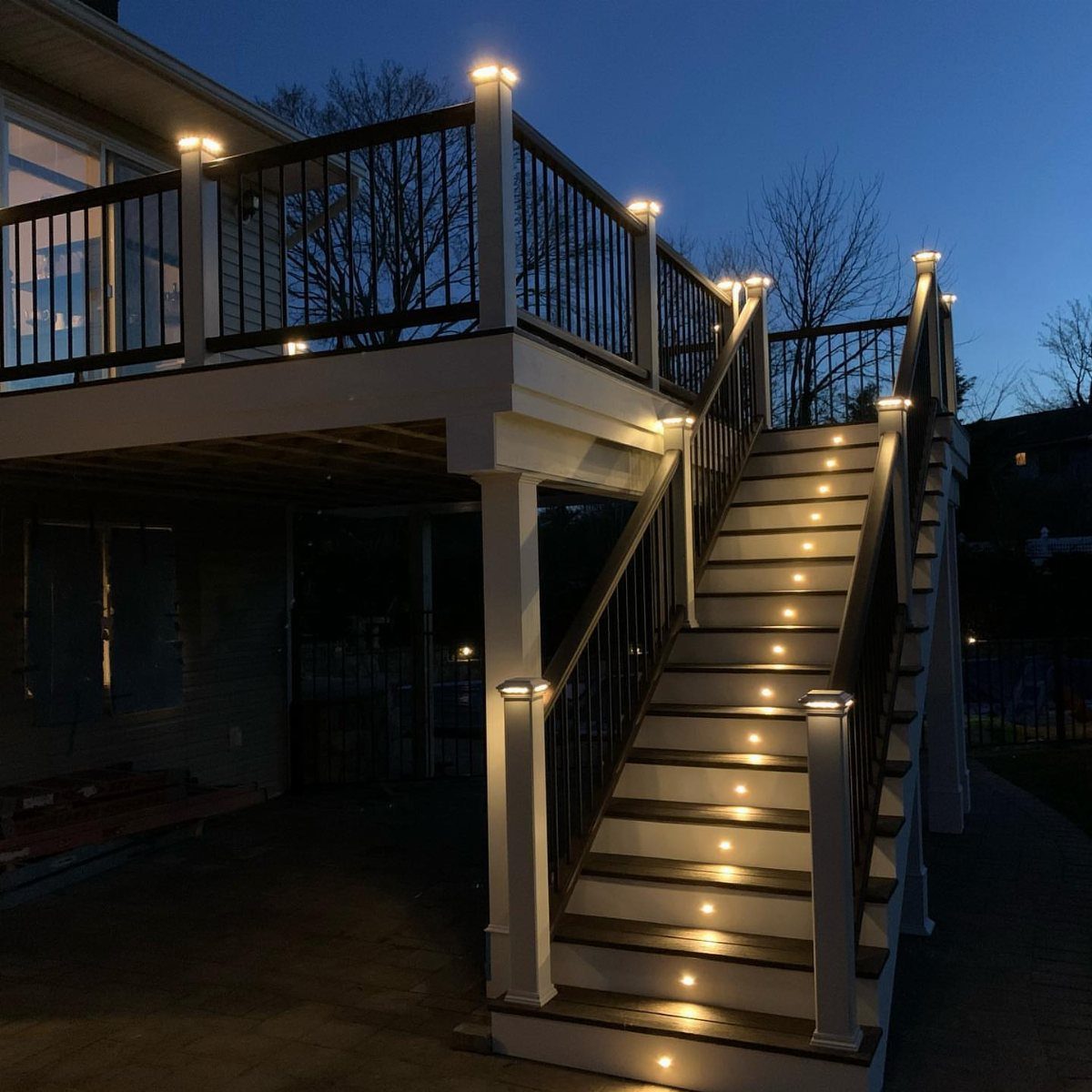 A lit two-story wooden deck with white posts and black railings at twilight.
