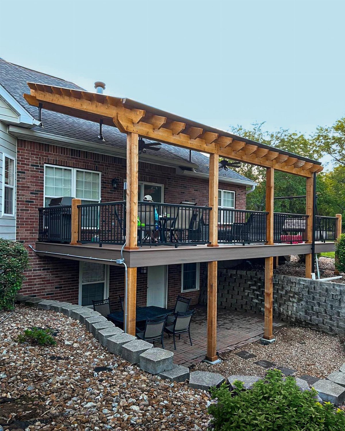 A large wooden second-story deck with black railings and a pergola structure attached to a brick house.