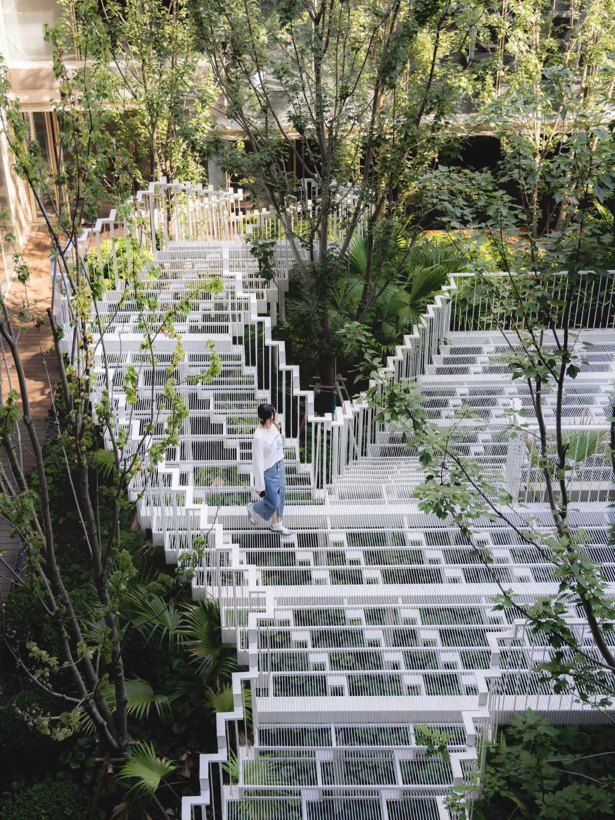 White geometric metal walkways winding through a lush green urban courtyard with many trees.