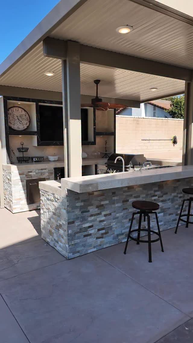 A stone-tiled outdoor kitchen island with a built-in grill and bar stools under a patio.