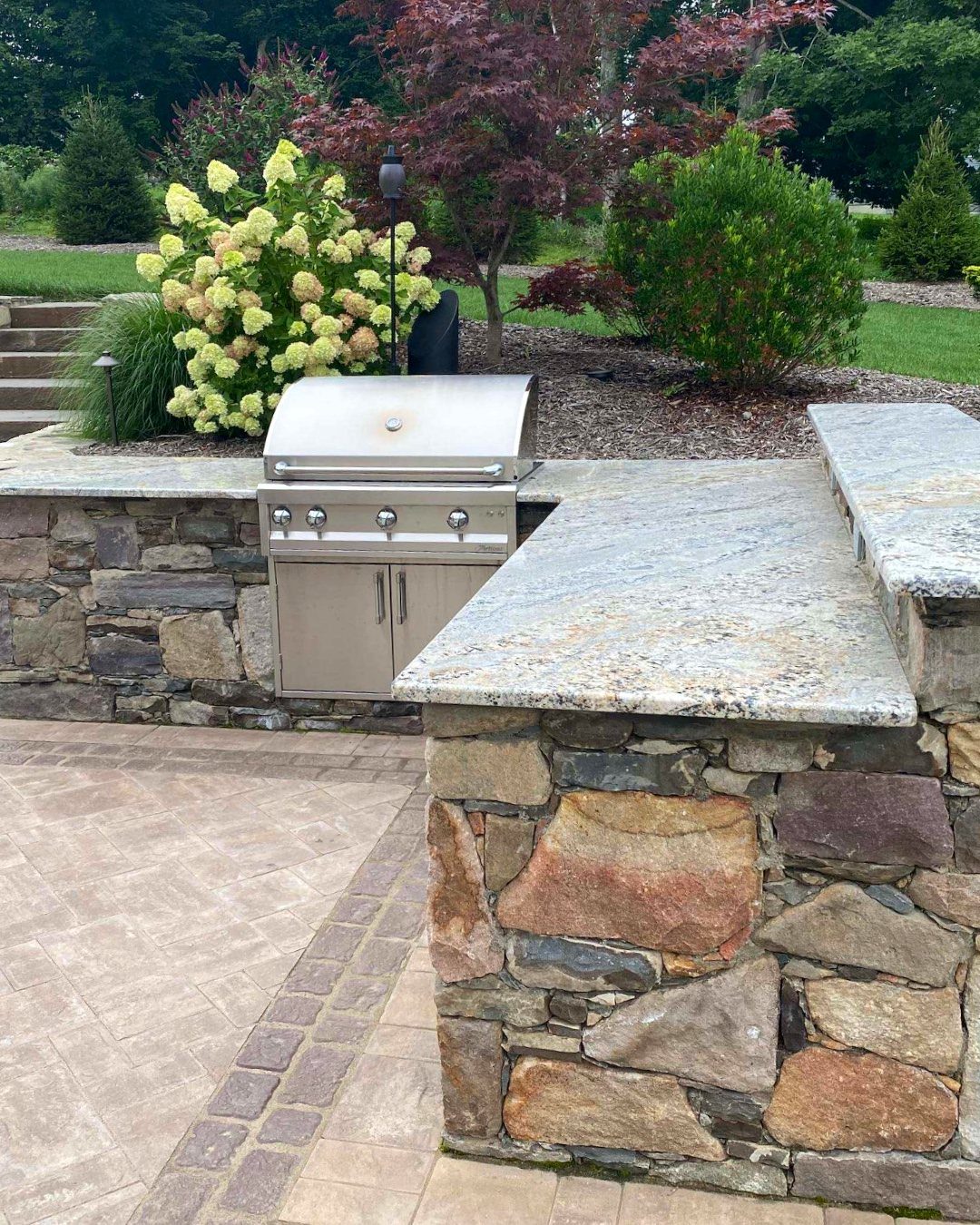 Stainless steel grill built into a stone outdoor kitchen counter surrounded by green plants and flowers.