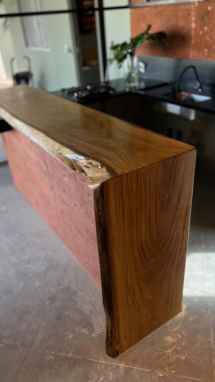 A thick wooden bar island with a live edge top and a reddish base in a kitchen.