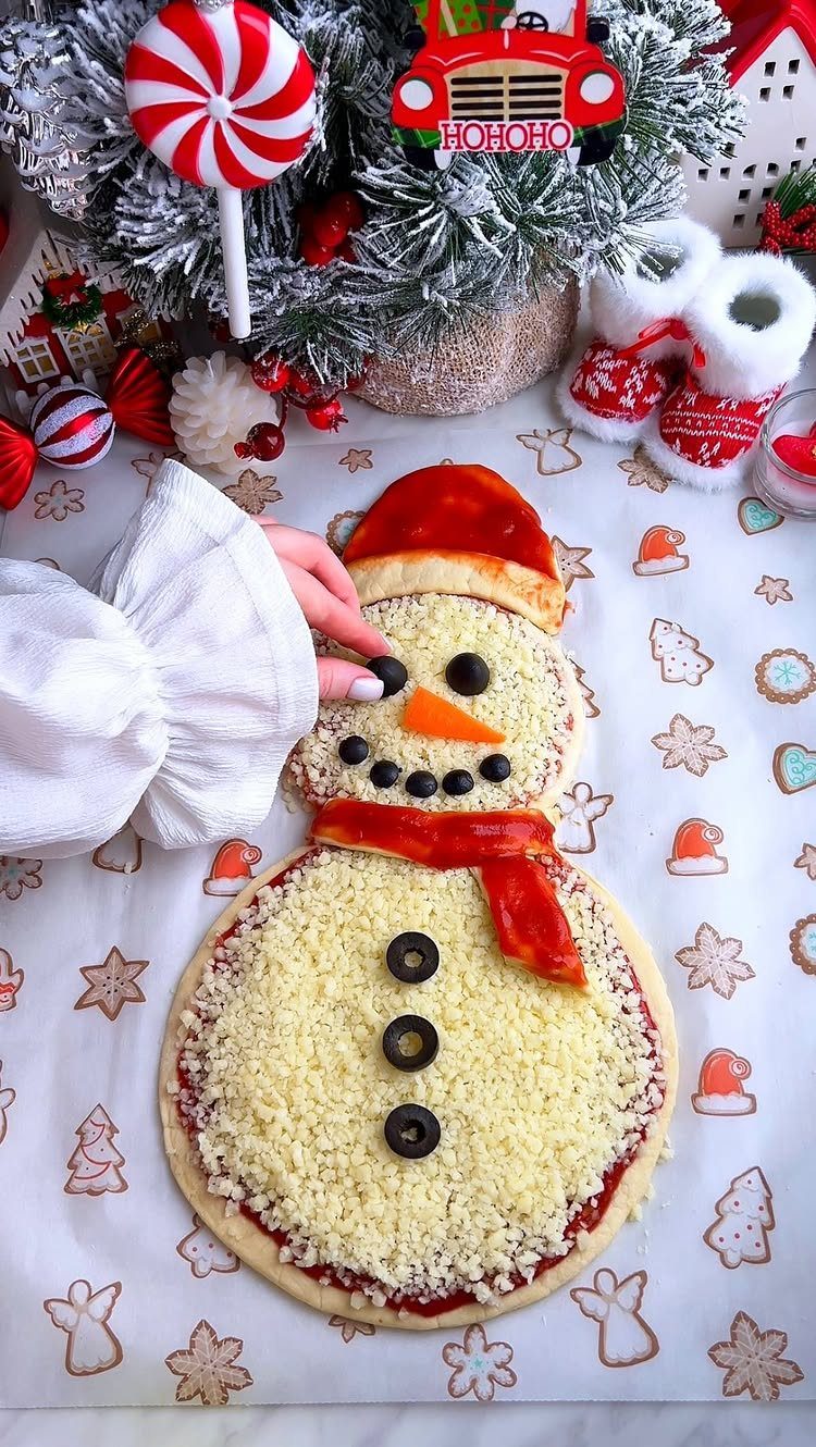 32 Easy Christmas Party Ideas for Food, Drinks, and Hosting in 2026 16 A snowman-shaped pizza decorated with olives and peppers on a festive holiday tablecloth.