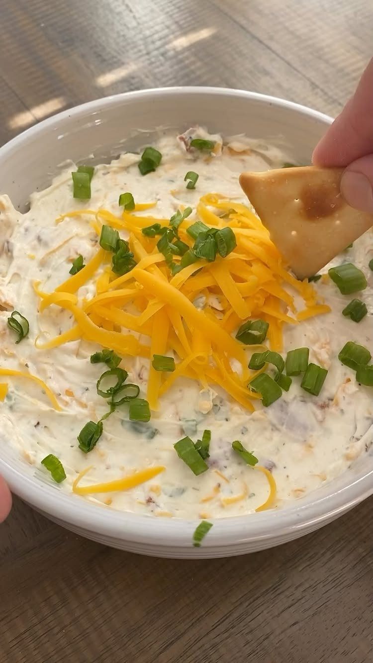 32 Easy Christmas Party Ideas for Food, Drinks, and Hosting in 2026 17 A hand dipping a pita chip into a bowl of cream cheese ranch dip topped with cheese and onions.