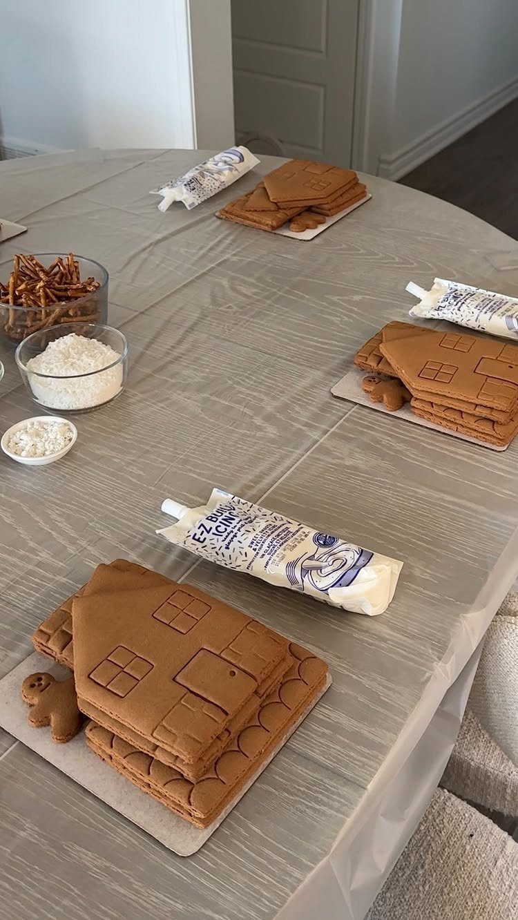 32 Easy Christmas Party Ideas for Food, Drinks, and Hosting in 2026 13 Gingerbread house kits with icing tubes and small gingerbread men on a grey tablecloth.