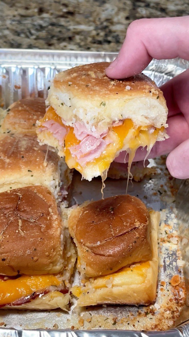 32 Easy Christmas Party Ideas for Food, Drinks, and Hosting in 2026 18 A hand lifting a warm, cheesy ham and cheese slider from a foil baking tray.