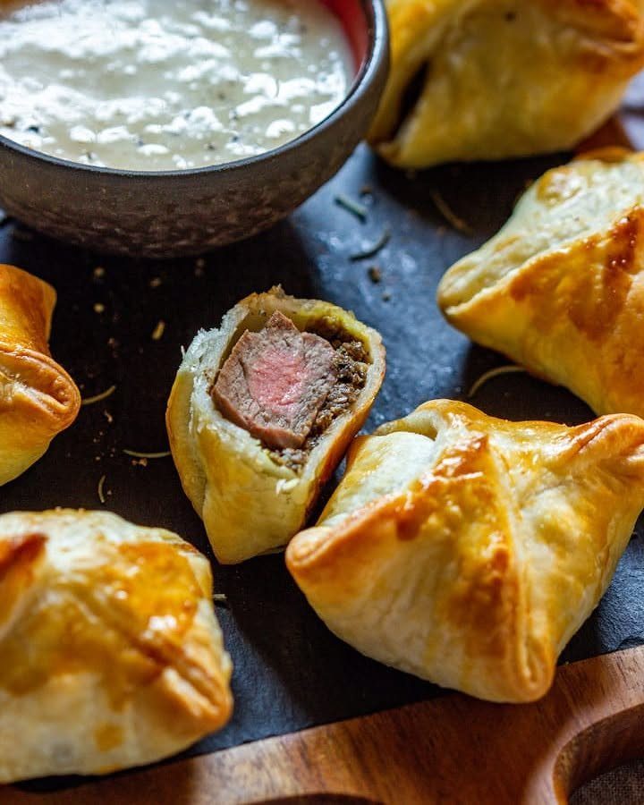 32 Easy Christmas Party Ideas for Food, Drinks, and Hosting in 2026 6 Mini beef wellingtons with golden puff pastry and pink beef filling on a dark slate board.