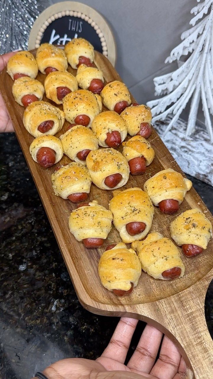 32 Easy Christmas Party Ideas for Food, Drinks, and Hosting in 2026 8 Pigs in a blanket appetizers arranged on a wooden serving board held by hands.