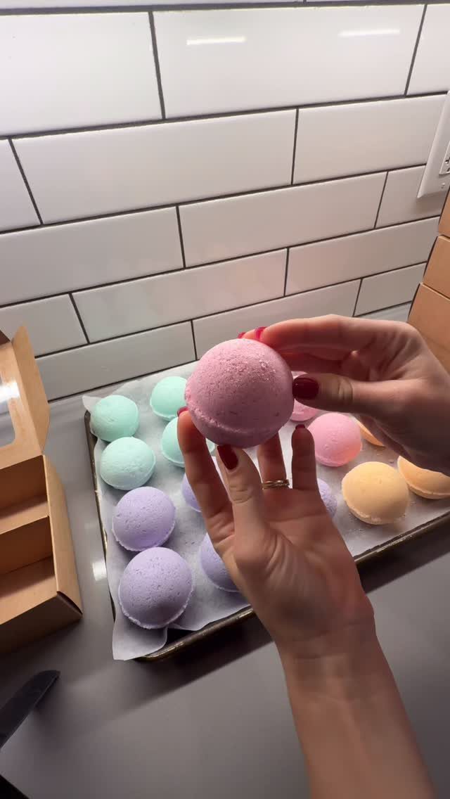 Hands holding a pink bath bomb above a tray of colorful pastel bath bombs.