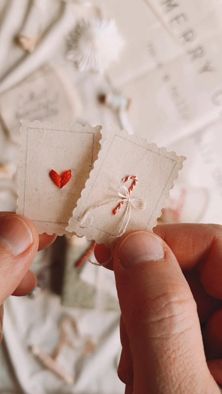 Hands holding two small scalloped paper tags with red embroidered heart and candy cane designs.