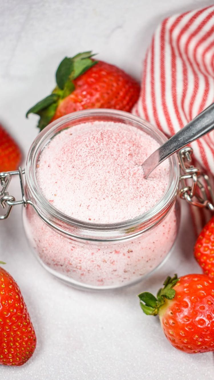 A glass jar filled with pink strawberry flavored sugar surrounded by fresh strawberries.