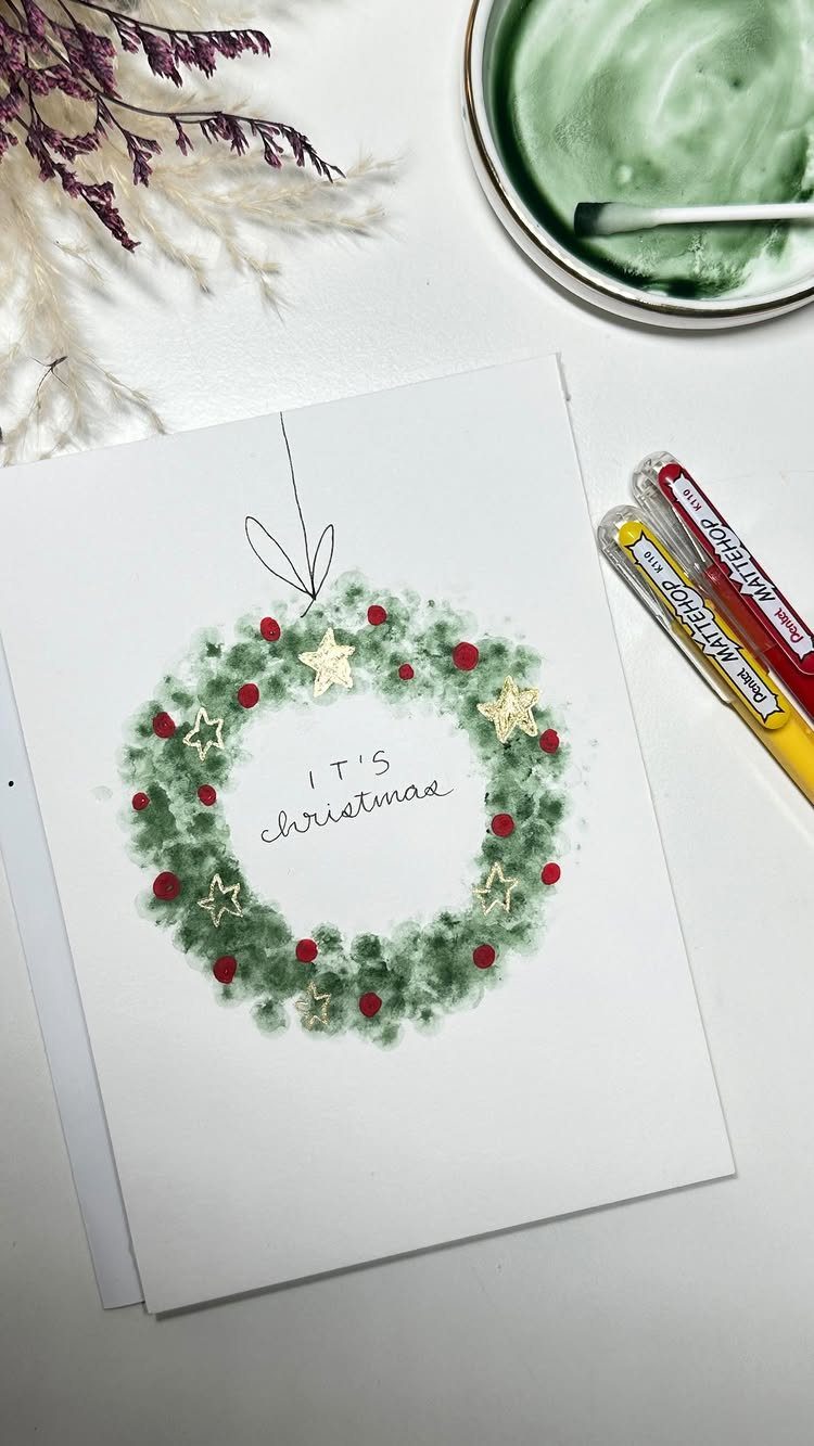 Hand-painted Christmas wreath card on a white table with art supplies and green paint.