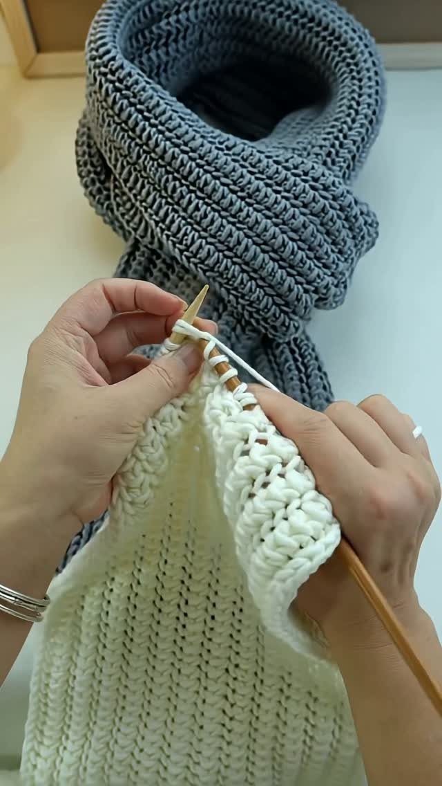 Hands knitting white yarn with wooden needles next to a finished grey scarf.