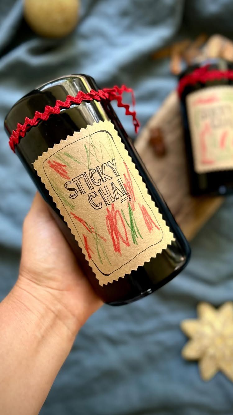A hand holding a dark glass jar labeled Sticky Chai with red ribbon decoration.