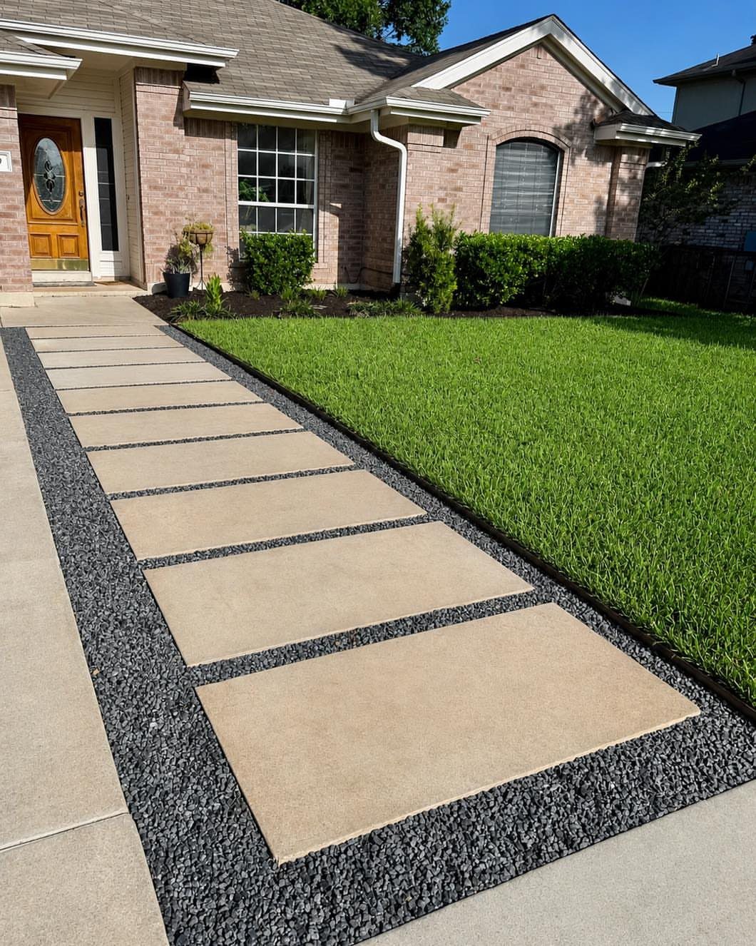 Rectangular concrete pavers set within a dark grey gravel pathway leading to a brick house.