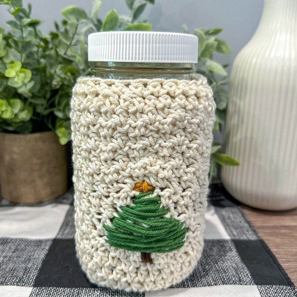 A mason jar wrapped in a cream crochet sleeve with a green Christmas tree design.