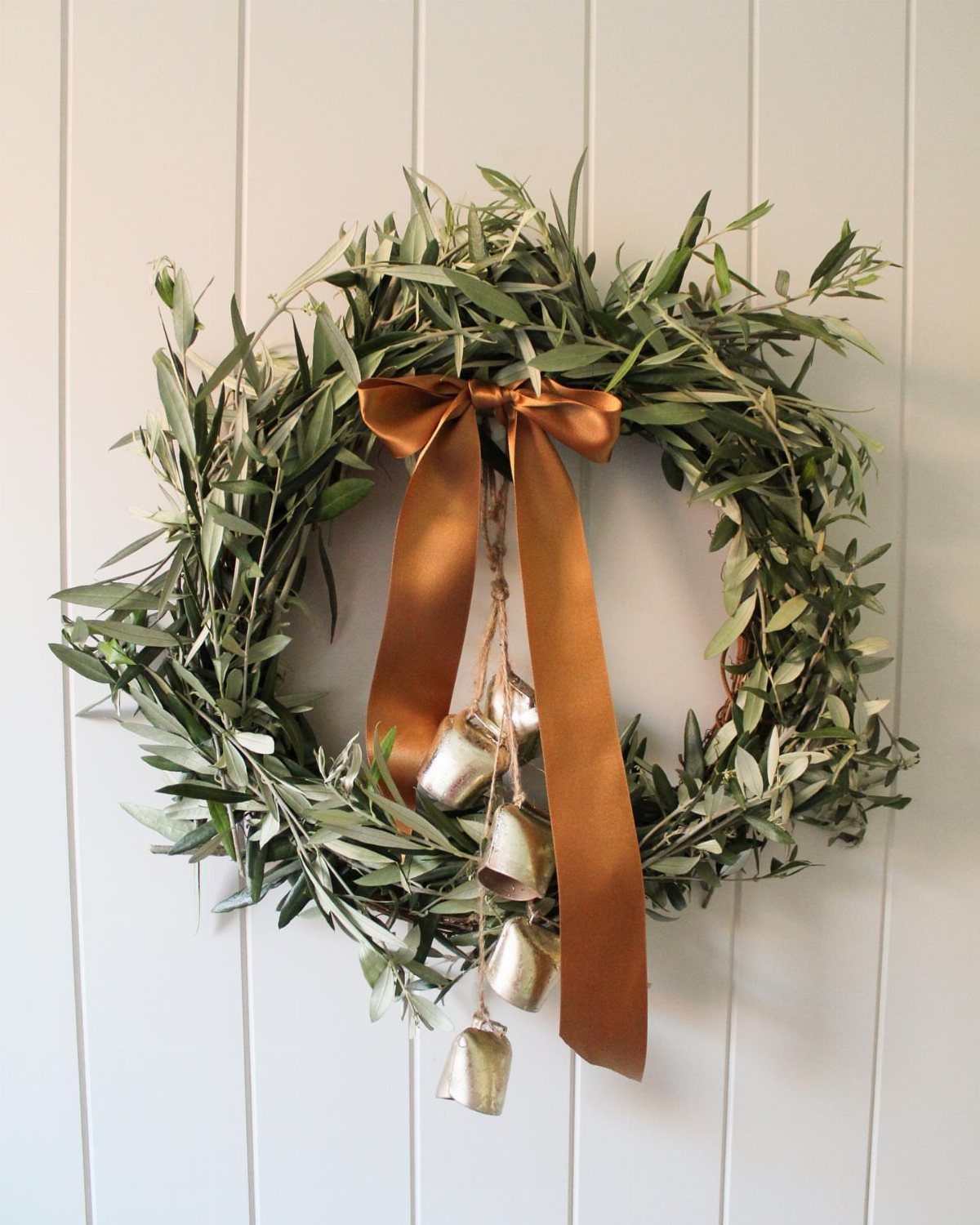 An olive leaf wreath with a copper ribbon and hanging gold bells on a white wall.