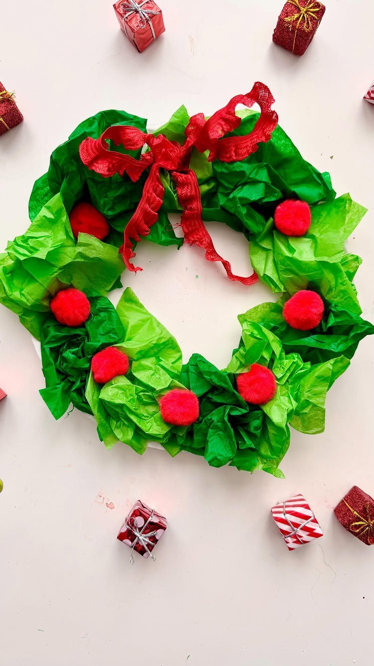 A green tissue paper wreath decorated with red pom-poms and a red bow on a white background.